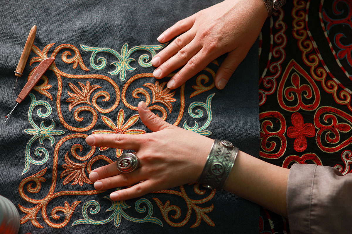 Close-up on two hands touching raised needlework in turquoise and orange on a dark gray textile background.