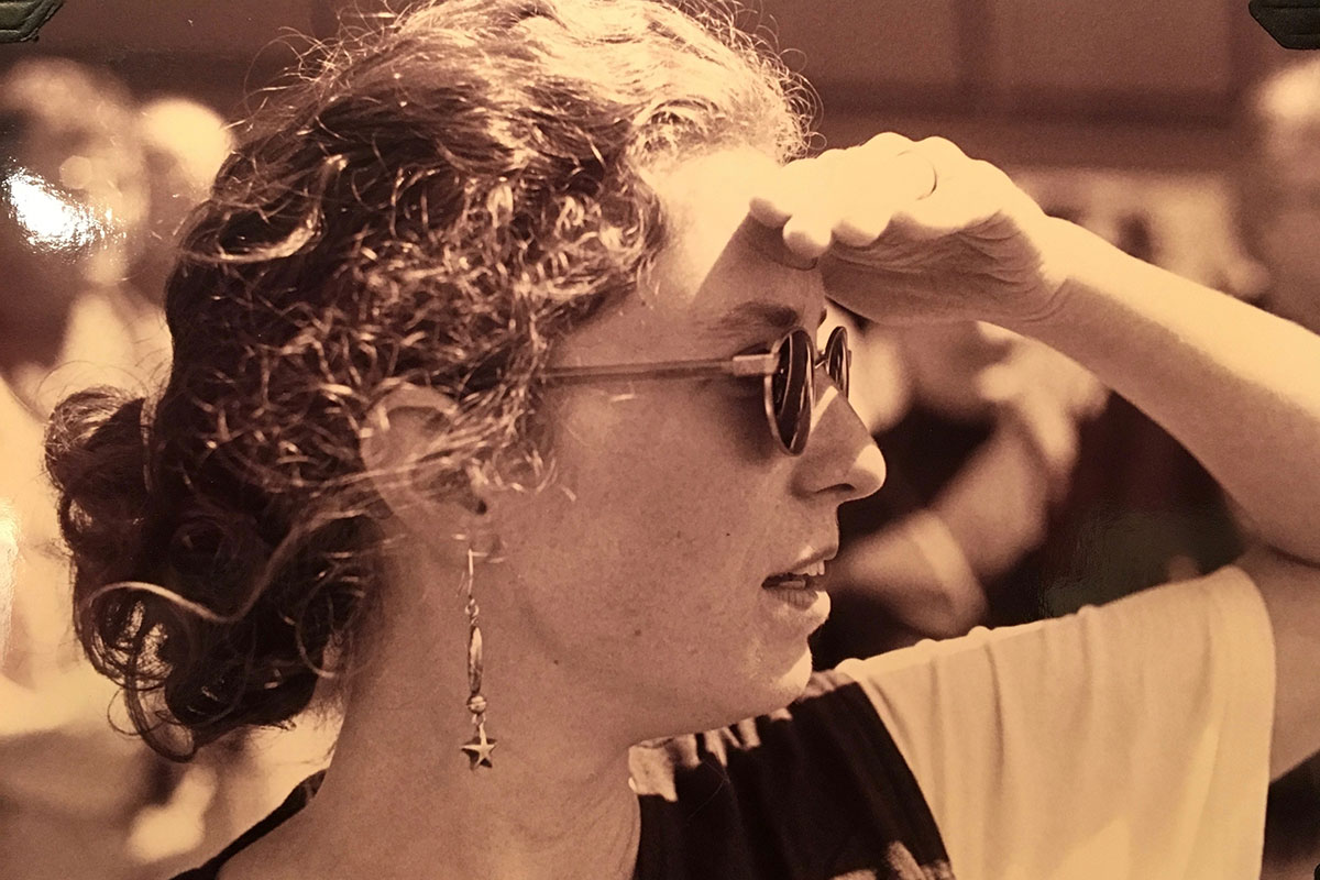 A woman wearing sunglasses and curly hair in a low bun looks into the distance, shading her eyes with one hand. Black-and-white photo.