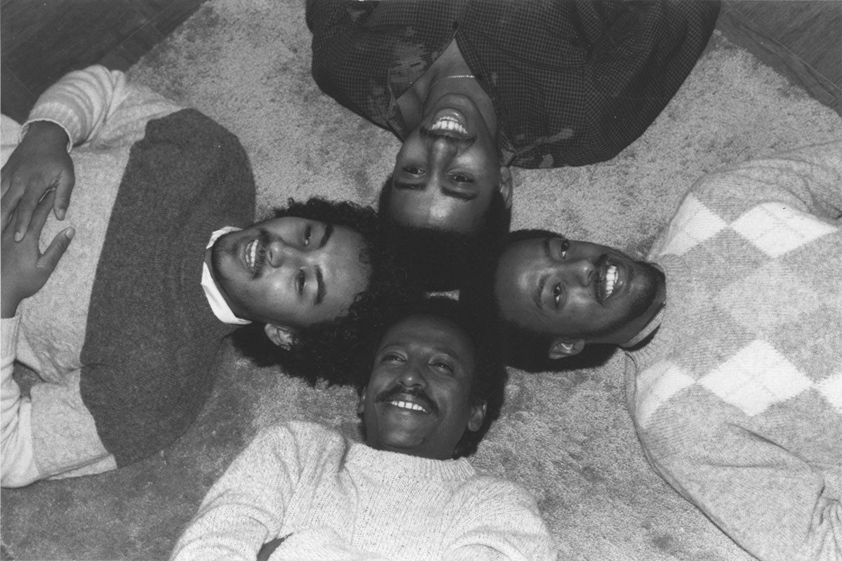 Four young men, smiling men lay on the ground, their heads together. Black-and-white photo.