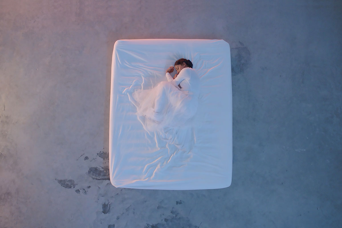 From above, a woman dressed in white, curled in fetal position, under a white sheet on a white mattress on a concrete floor. 