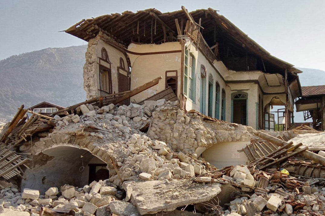 Some walls and parts of a roof remain of a house after an earthquake, with a pile of rubble at the base.