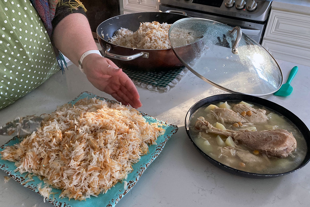 Closeup on a person's hand gesturing toward a square, turquoise plate full of fried rice. Beside it is a bowl of soup with a turkey drumstick and vegetables. Behind them is a pot containing more rice.