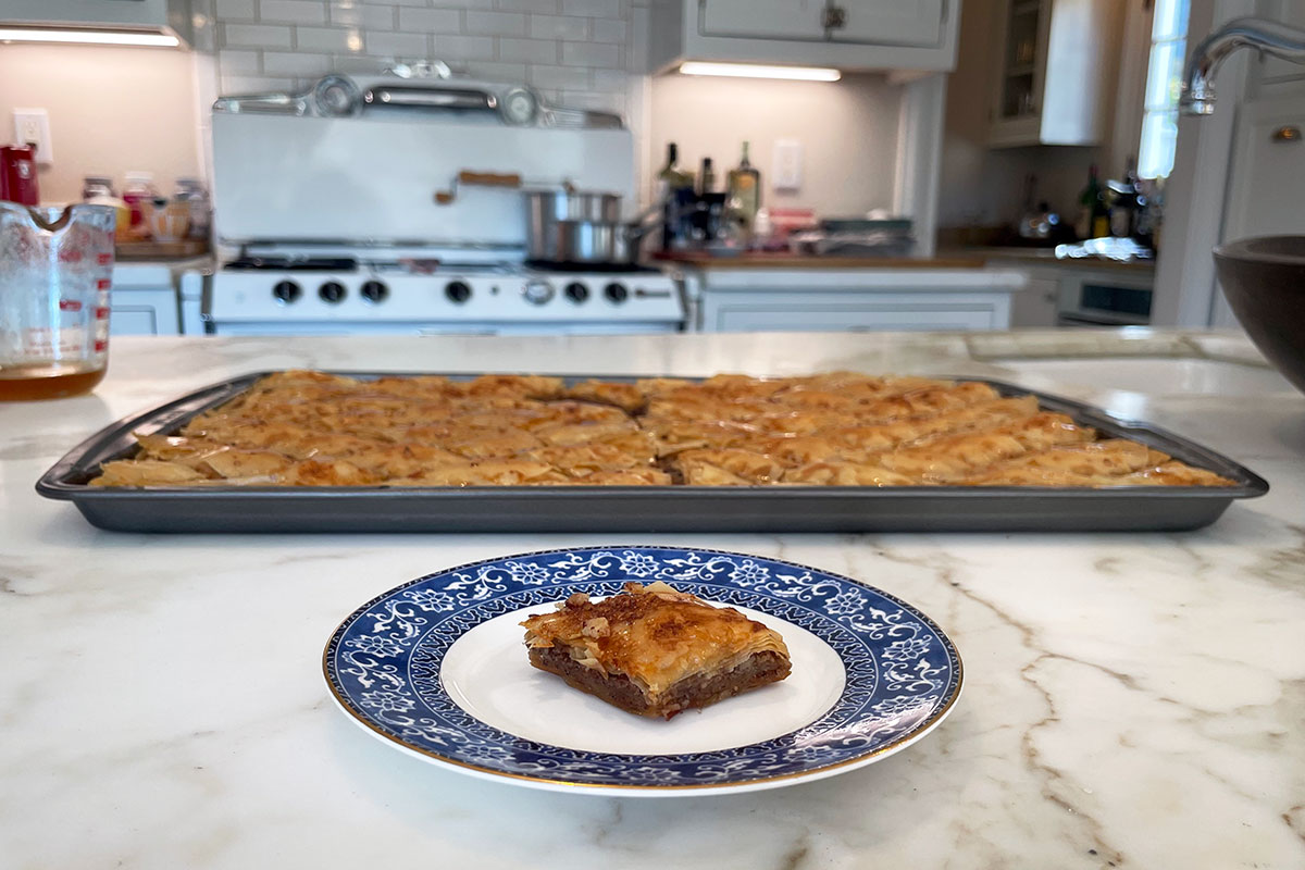 On a white marble kitchen counter, a plate with a single piece of flaky baklava and a baking tray full of the rest of the baklava.