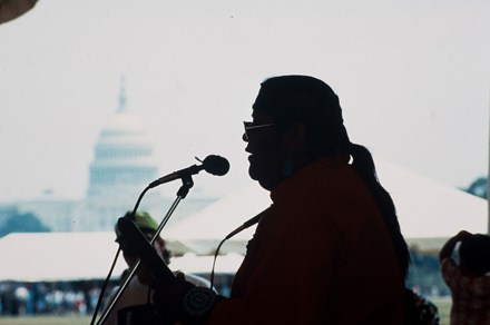 A silhouette of a man with a ponytail and aviator glasses faces left, against the backdrop of the U.S. Capitol Building. He leans into a microphone and holds a musical instrument.