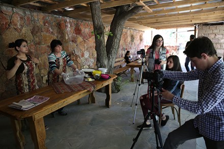 On the right, two young women and a young man sit with video cameras and tripods filming two women wearing traditional Armenia dress on the right.