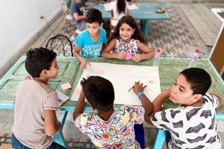 Five children sit together around a pale-green table, holding pencils. In the middle of the table is a large, blank sheet of paper with pink sticky notes on it--ready to be drawn on.