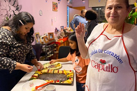 In the foreground, a woman wearing a white apron with a Spanish-language slogan smiles at the camera. Behind her, a young woman and young girl places strips of brightly colored fruit candy and nuts on top of a rectangular ring cake. 