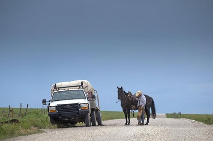 A man in cowboy hat stands in the middle of a rural road next to a black horse and white pickup truck.
