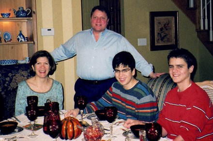 A family of four at an elaborately set dinner table: mom and two brothers seated, and dad standing between them.