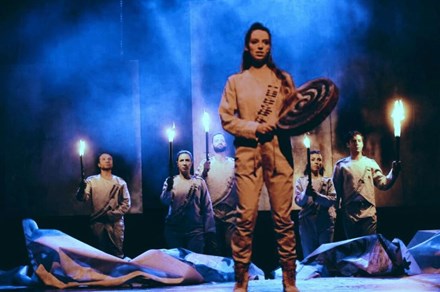 A woman stands on stage, holding a hand drum and a beater stick. Five actors behind her hold up torches. They all wearing matching white outfits with mock bandoliers. The stage is foggy and lit blue.