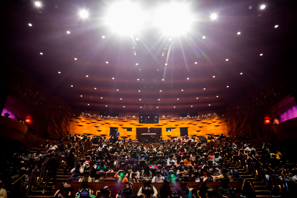 An auditorium is pictured facing the audience. The room, with orange walls and a purple ceiling, is packed with people wearing black, virtual reality headsets sitting in their chairs.