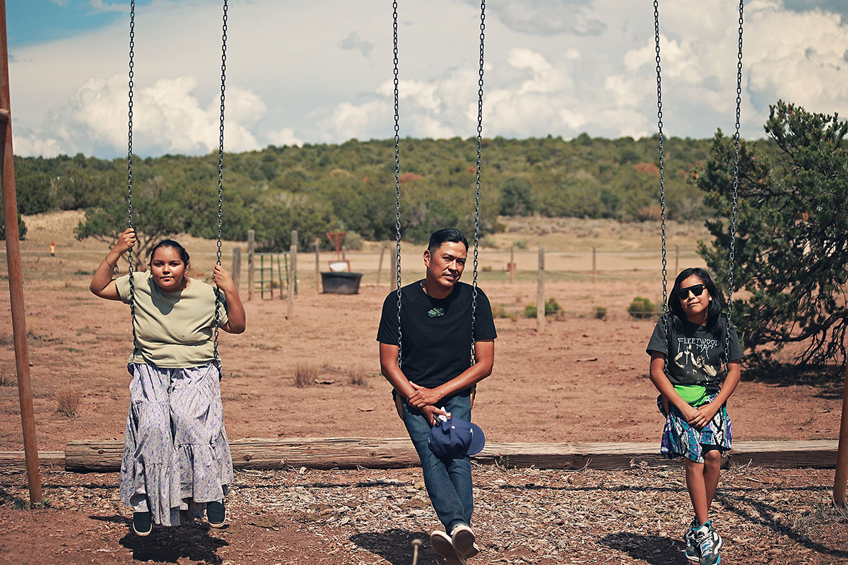 Two children and an adult sit on three swings in a dry landscape with trees in the background.