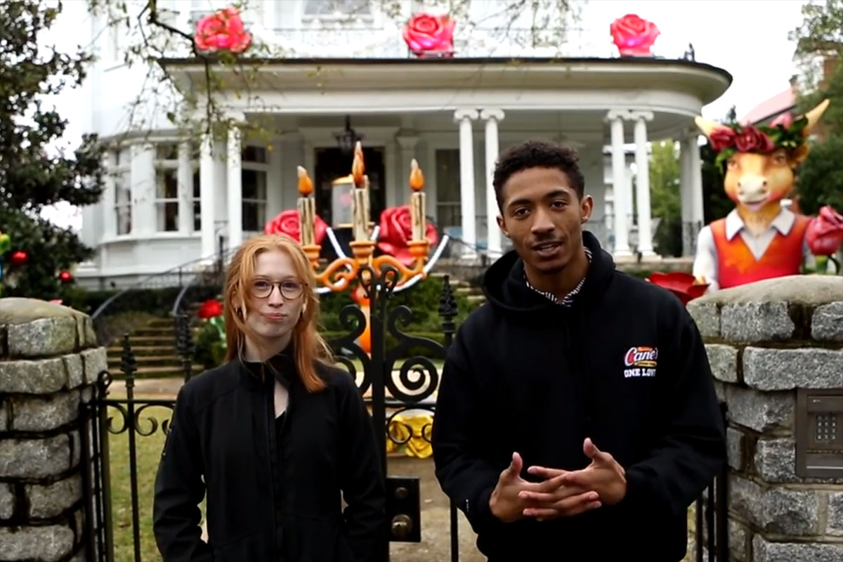Two young adults face the camera and talk in front of a gate and a decorated large house.