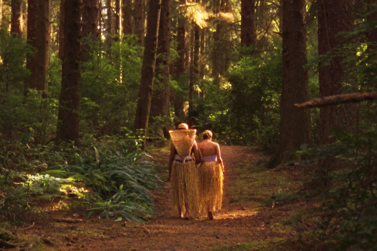 Two people in light-colored grass skirts, one carrying a woven basket on their back, walk down a forest path, dappled with sunlight.